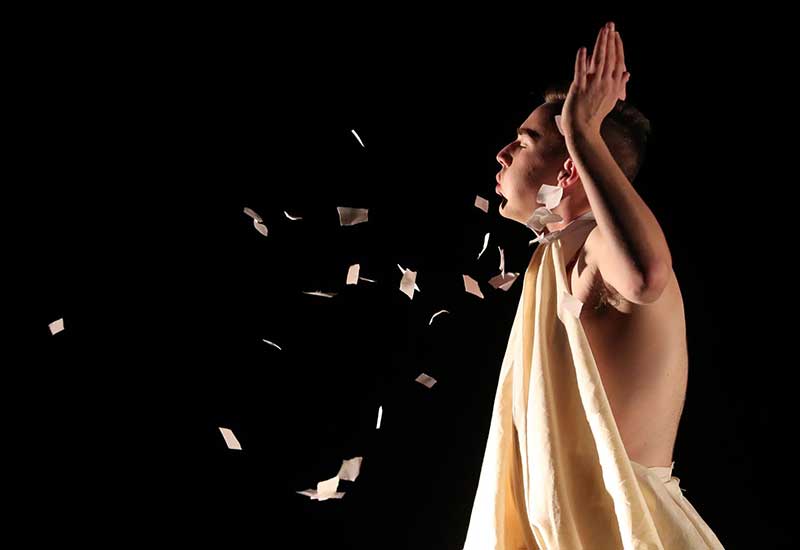 a performer wearing a toga blows scraps of paper across the air during a dance performance