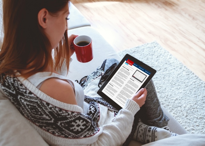 A woman sitting on a couch reading on a tablet and holding a coffee mug.