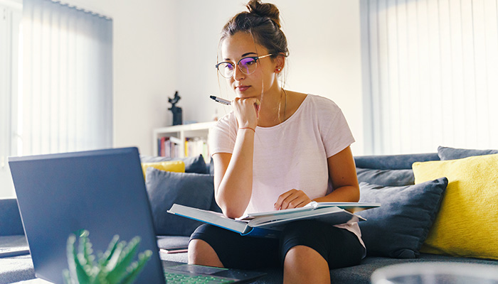 a woman sits on a couch holding a book. she is looking at a laptop on the table in front of her