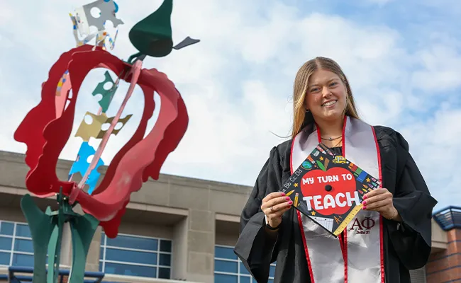 a student in graduation regalia shows off her mortar board bearing the words my turn to teach