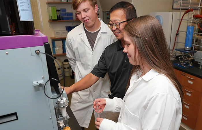 an instructor demonstrates lab equipment to two students in lab coats