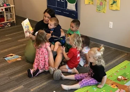 Teacher reading a picture book to a small group of toddlers seated on the floor in a classroom.