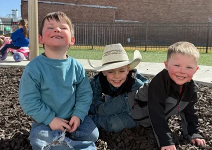 Three young boys smiling while sitting on playground mulch, one wearing a cowboy hat.
