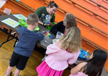 Children gathered around a table, working on colorful crafts with an adult supervisor in a gymnasium setting.