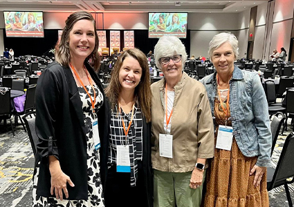 Four women wearing conference badges pose together in a large event hall with round tables and presentation screens in the background.