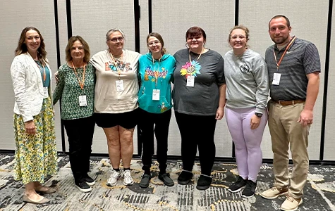 Seven adults standing in a line at a conference, smiling and wearing name badges.