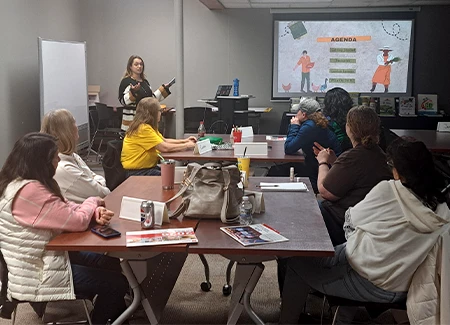 Presenter leading a workshop in a conference room while attendees sit at tables and watch an agenda slide on a projector screen.