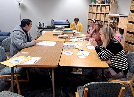 Small group meeting around a conference table with papers and books spread out, in an office with shelves in the background.