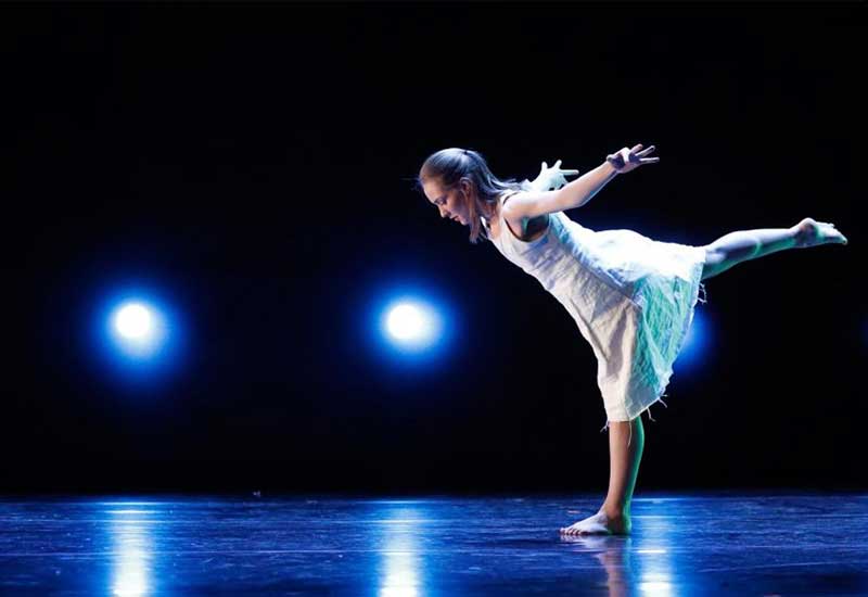 a student performing a dance piece leans out in front of a black background decorated with blue lights