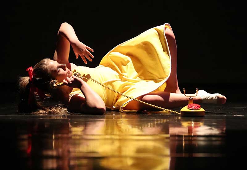 a performer in a yellow dress lies on the stage holding a phone during a dance concert