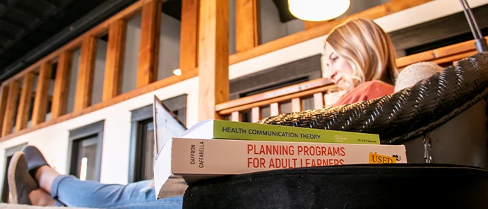 a student lounges while working at her laptop, a pile of communication textbooks sits in the foreground