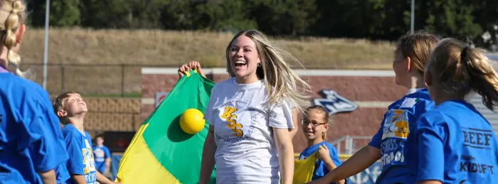 Smiling woman in a white T-shirt runs alongside children in blue shirts during an outdoor school activity, with a colorful play parachute in the foreground.
