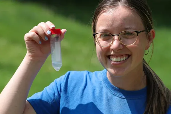 a student stands in a field and holds up a sample vial