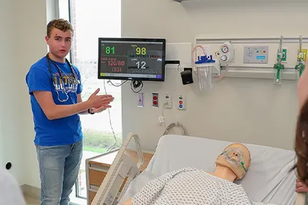 a student stands next to a medical dummy and points at a monitor