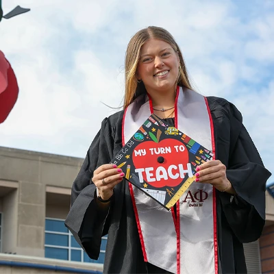 a student in graduation robes stands outside the coe building. her cap has been decorated with the words my turn to teach