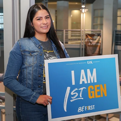 a student stands in the college of education building with a sign that says I am First Gen