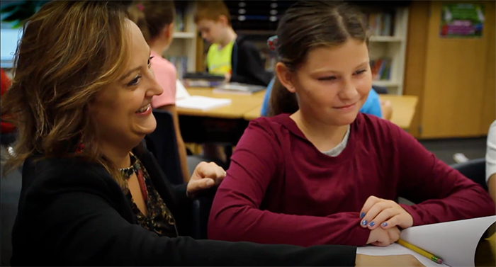 Female teacher kneeling beside female student's desk to assist her with an assignment.