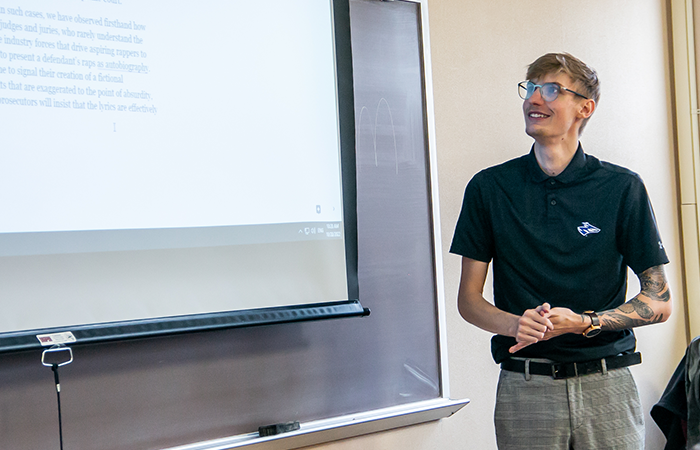 A young man in a black polo shirt presents information in front of a projector screen during a classroom setting.