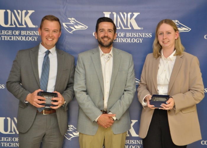 Two students holding awards with professor in front of backdrop. 