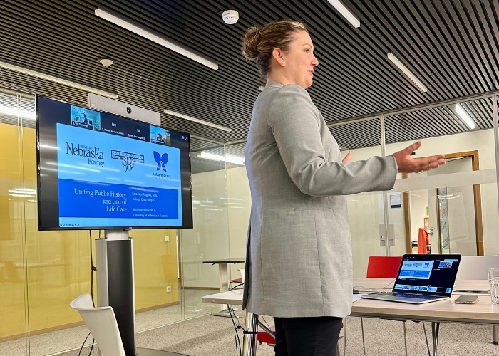 A side view of a woman wearing black slacks and a grey blazer giving a presentation with a slideshow being shown on a TV screen behind her. There is a table in front of her with an open laptop on it and she has her hands out in front of her in a gesture.