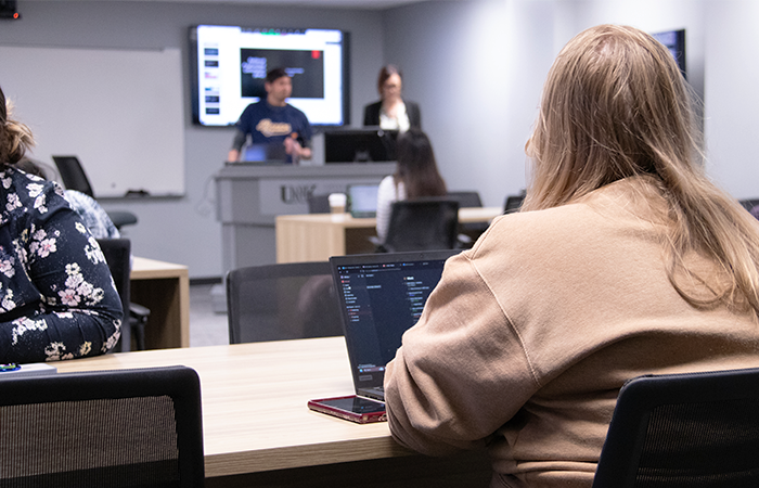Students engaged in a classroom setting, with two people presenting at the front and others using laptops.