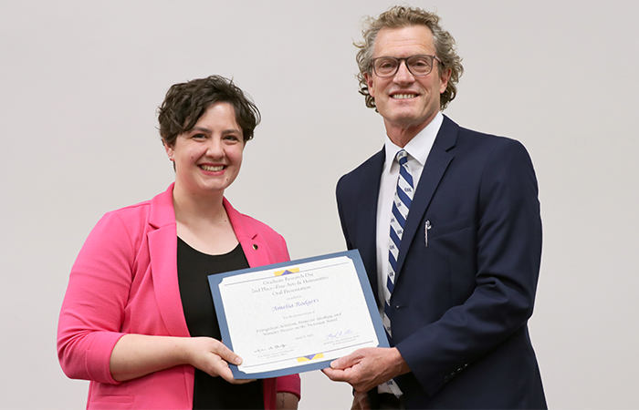 A woman in a pink blazer stands next to a man in a suit, both smiling as they hold up a certificate.