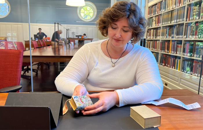 A woman examines a small decorative book in Harvard's library, surrounded by shelves filled with books.