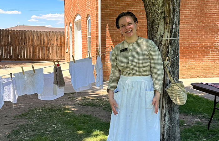 Katie Henry in historical reenacting clothing stands beside a tree, with clothes hanging on a line behind her. The setting reflects a 19th-century outdoor scene.