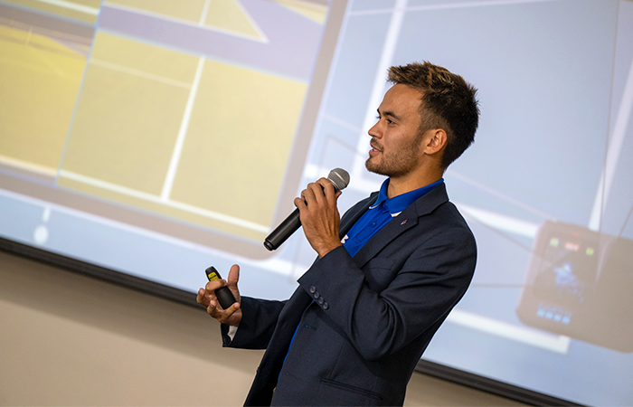Aaron Osmond wearing a blue suit delivers a presentation, holding a microphone and a remote device, with a digital display behind him.