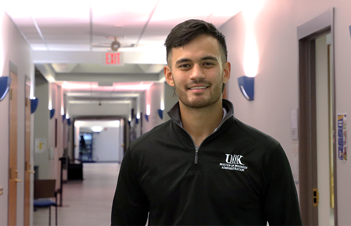 A man with dark hair stands in a bright hallway, wearing a black quarter-zip pullover embroidered with “UNK Master of Business Administration.”