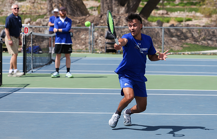 A male tennis player in a blue UNK uniform reaches for a tennis ball while playing on an outdoor court.