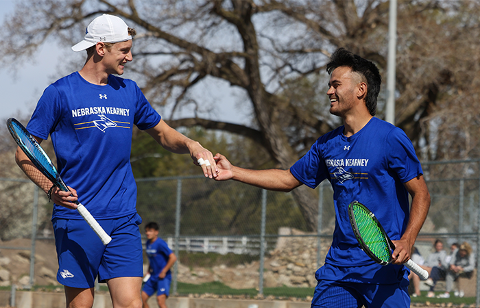 Two UNK mens tennis players celebrate a successful play with a high-five on the court.