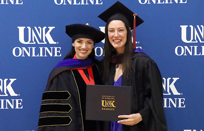 Two women celebrate at a graduation ceremony, one in academic regalia holding a diploma, with a backdrop featuring the UNK Online logo.
