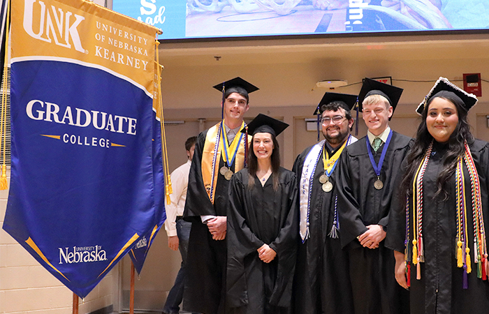 Graduates posing in their caps and gowns with a University of Nebraska Kearney Graduate College banner.