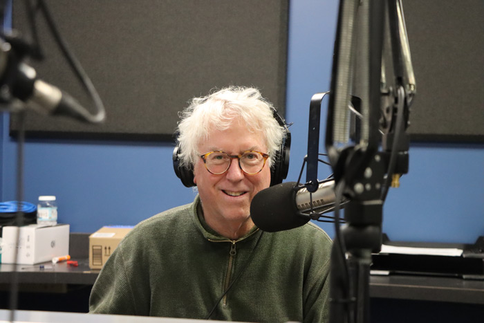 Rick Brown smiles with headphones on seated behind the microphone in a radio studio.
