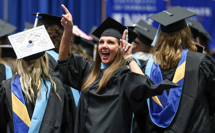 Woman wearing UNK graduation regalia pointing up with a smile.