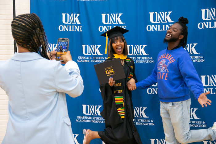 Woman takes a photo on a cellphone of a woman in UNK graduation regalia holding hands with a man, both have their foot popped up in the air in front of a UNK Online backdrop.