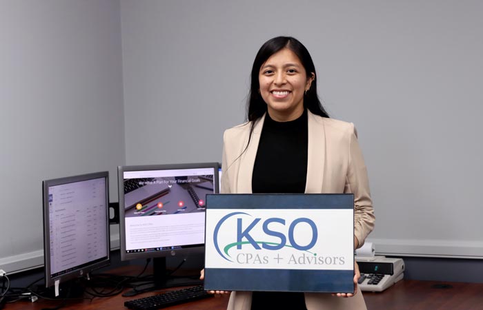 A woman in a beige blazer holding a sign for KSO CPAs + Advisors, standing in an office with computer monitors in the background.