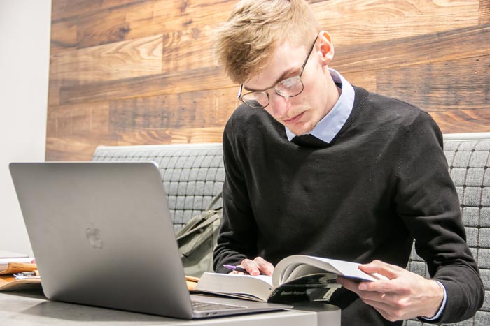 A young man wearing glasses is reading a book while sitting at a table with a laptop open in front of him.
