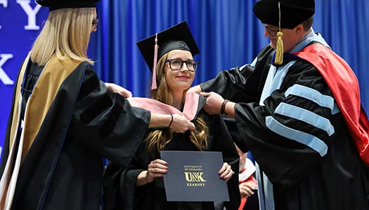Woman in glasses being hooded at UNK's commencement ceremony.
