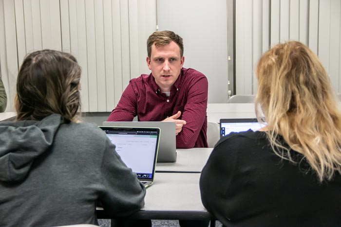 Man and two woman seated at a table in a classroom setting - all have laptops in front of them.