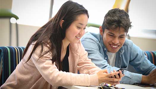 Two students sitting at a table, smiling and looking at a phone