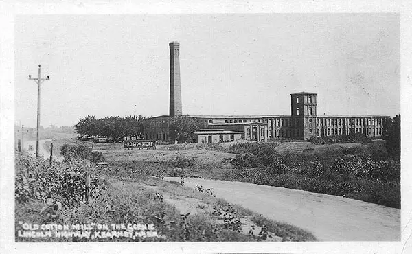 Black-and-white historic photo of a cotton mill complex with a tall smokestack beside a rural road.
