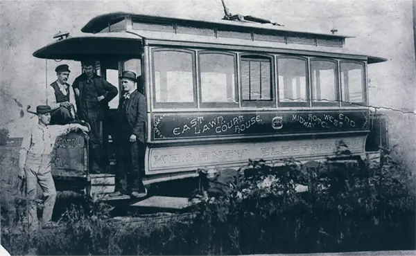 Vintage black-and-white photo of an early electric streetcar with several men standing on the platform beside it.