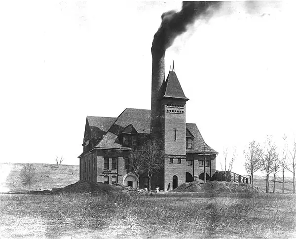 Historic industrial building with a tall smokestack emitting dark smoke in a rural landscape.