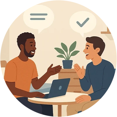 Two men engaged in a friendly conversation while seated at a table, with a laptop open in front of them and a plant in the background.