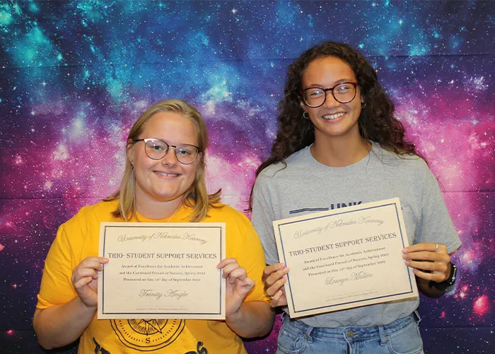 two students hold up certificates in front of a cosmic space background