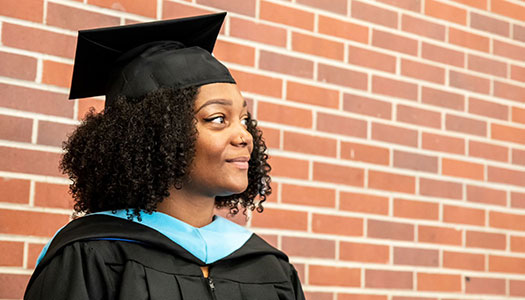 a woman in a graduation cap and gown stands in front of a brick wall