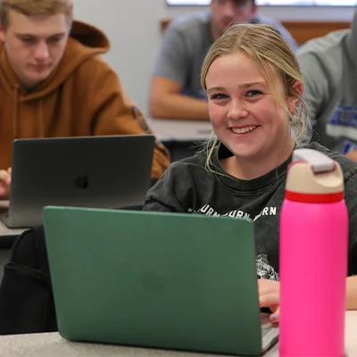 a student sitting in front of a laptop smiles at the camera