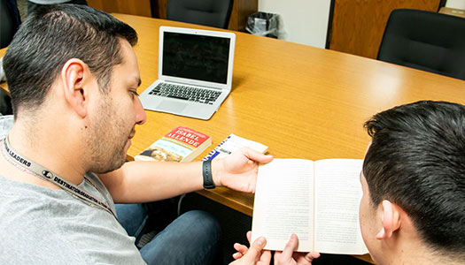a student and instructor sit at a table with a laptop looking at a book